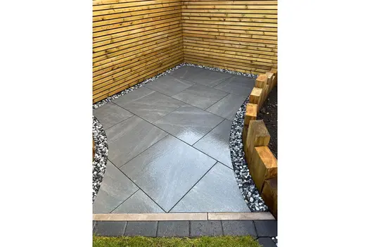 A modern outdoor space featuring Allora Nero 600x600 porcelain paving, bordered by a wooden slatted fence and gravel edging.