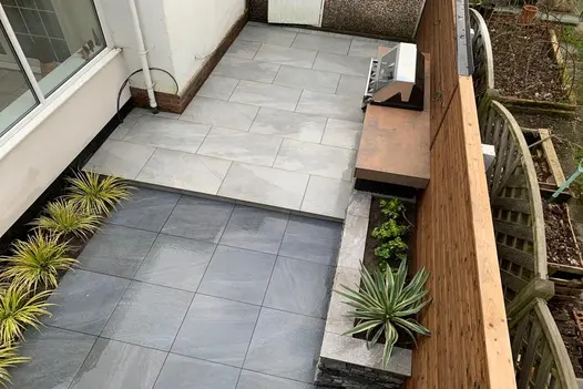 A bird's-eye view of a split-level outdoor space featuring contrasting porcelain paving, a barbecue area, and a wooden fence.