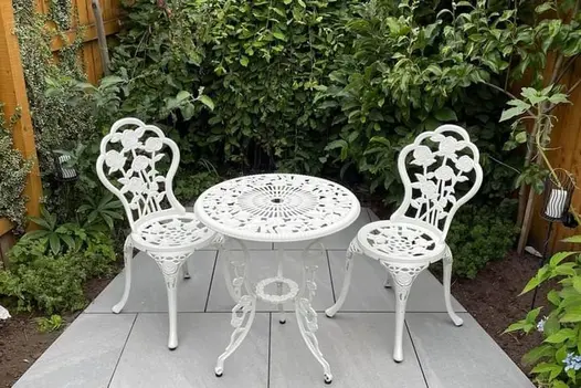 A charming patio paved with Allora Silver Porcelain, featuring a white metal bistro set surrounded by lush greenery.