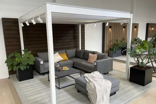 In indoor showroom, Proteus White aluminium pergola, with planters and furniture beneath, sits on composite deck.