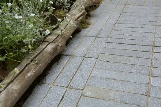 Antique Grey Limestone Brick Pavers laid in a neat linear pattern beside a rustic driftwood garden border and soft white planting, showcasing their cool grey tones and textured finish.