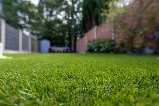 A close-up shot of St Tropez artificial grass with a blurred garden fence in the background.