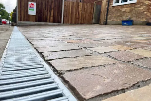 Raj Green Sandstone Setts used on a driveway, showing their natural green-brown colour variations and tumbled edges alongside a metal drainage channel.
