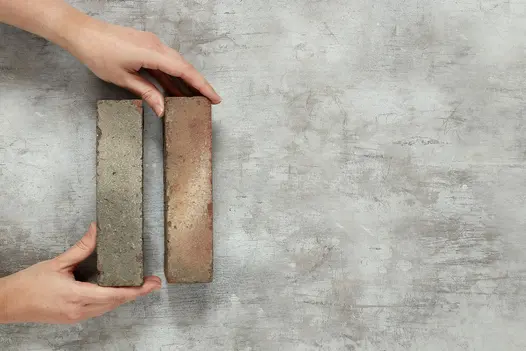 2 hands place 2 Bexhill clay paving bricks, in red and grey with white markings, next to each other on pale background. 
