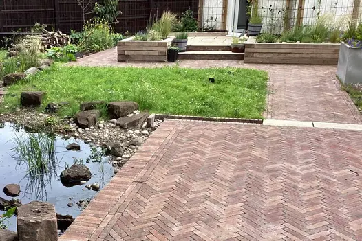 Bexhill Clay Brick Pavers laid in a herringbone pattern, creating warm-toned garden pathways beside a natural pond and wild-style planting.