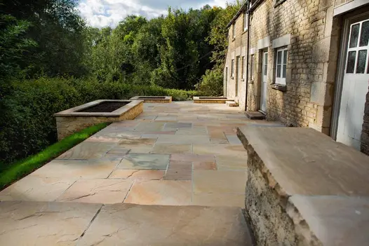Camel Dust Indian Sandstone paving laid in a traditional multi-size pattern beside a stone cottage, with raised planters and countryside views.