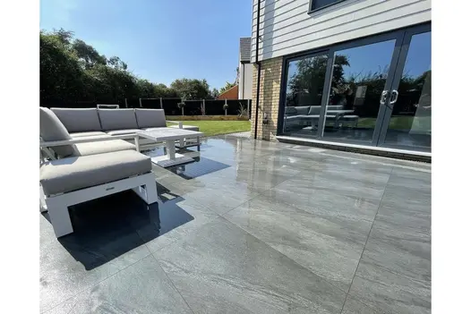 Spacious garden patio with grey porcelain tiles, white furniture, and freshly mown lawn bordered by dark fencing.