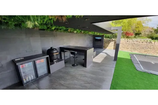 Outdoor kitchen with bar, BBQ, and grey paving, set beneath a pergola with overhead greenery and garden views.