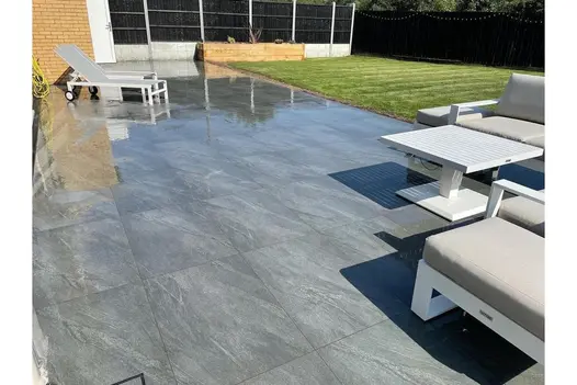 Wet-effect grey porcelain paving with white outdoor furniture in a modern garden beside a light-brick home.