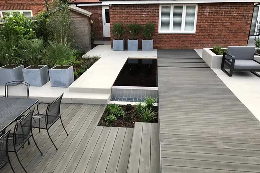 Charcoal DesignBoard Composite Decking used throughout a modern garden design, featuring grey seating, pale porcelain paving, raised planters, and a reflective sunken pond at the centre.