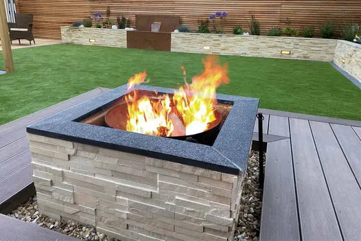 Chestnut Grooved Composite Decking installed around a modern garden firepit with stacked-stone cladding, artificial lawn, and contemporary raised planters.