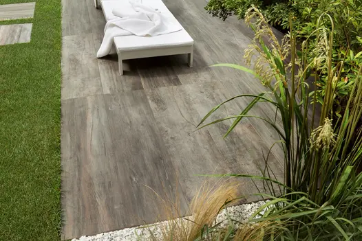 Cinder Porcelain paving with a natural wood-effect finish, used beside a lawn and planted borders with a sun lounger.