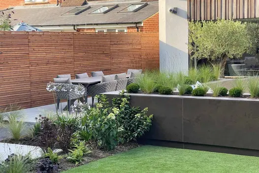 Dark Mocha Porcelain Cladding used to finish a sleek raised planter, paired with architectural grasses and a contemporary outdoor dining area for a modern garden design.