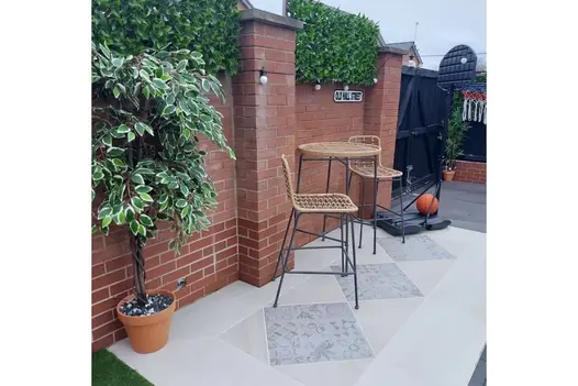 Outdoor seating area with feature Deco 600x600 Porcelain tiles inlayed in light paving, set against a red brick wall with faux greenery and lights.