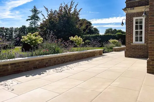 Florence Beige Porcelain paving creating a smooth, elegant terrace beside a brick house, with raised flowerbeds and hydrangeas in bright sunlight.