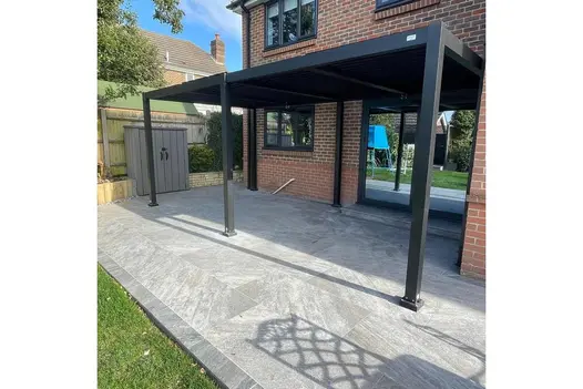Covered patio with Gaja Grey Porcelain Paving, extending from a red-brick house into a landscaped garden.
