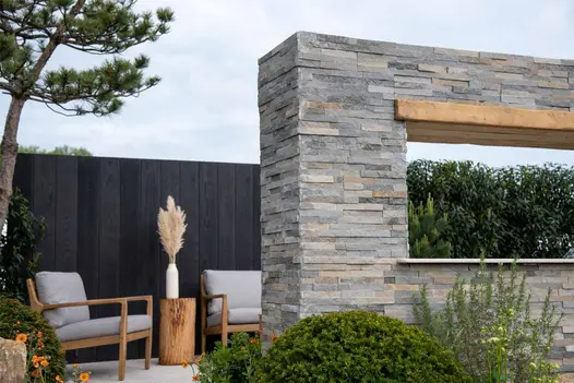 Graphite Sandstone Stone Cladding used to build a contemporary outdoor feature wall and column, showcasing layered grey tones beside a modern seating area in a landscaped garden.