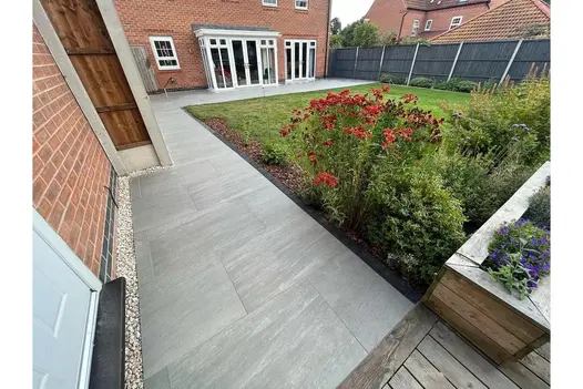 A sleek black fence frames this space, where Kandla Grey Porcelain Paving enhances the lush garden filled with red flowers.