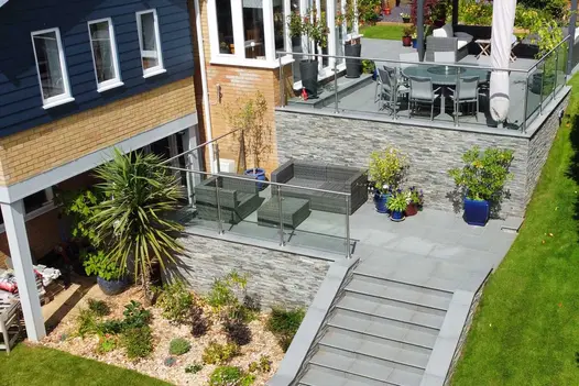 Kandla Grey Sandstone Stone Cladding used on raised terrace walls in a multi-level garden, adding texture and a natural stone finish alongside glass balustrades, seating areas, and landscaped planting.
