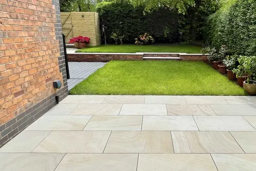 Light Buff Sandstone Paving forming a clean, contemporary patio overlooking a raised lawn, with smooth buff-toned slabs laid beside a brick house.