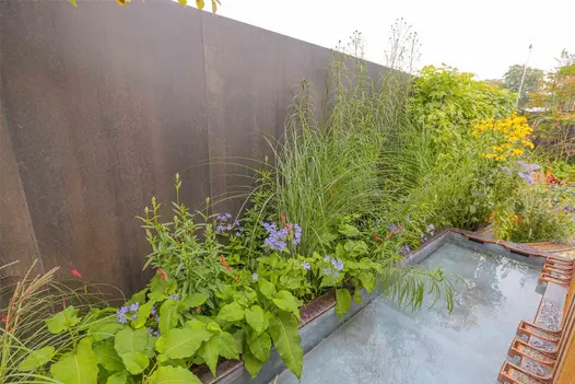 Metal Dark Porcelain Cladding used as a dramatic backdrop to lush planting and a reflective water feature, adding industrial depth and contrast.
