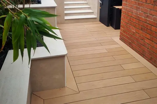 Mocha DesignBoard composite decking in a warm light-brown tone installed beside a red-brick wall and raised planters, leading towards porcelain-clad garden steps.