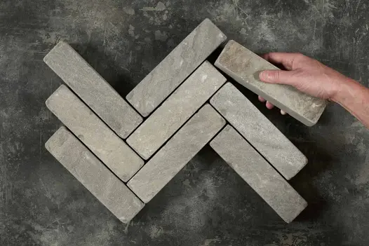 Hand places single Black sandstone patio brick against 8 laid herringbone on dark grey background. Free UK delivery available.