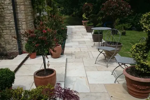 Tumbled Mint Sandstone Paving used to create a light, characterful garden pathway, surrounded by terracotta pots, clipped shrubs, and metal bistro chairs along a stone-built house.