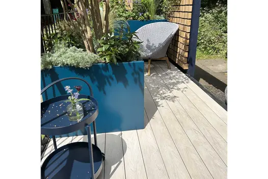 Polar DesignBoard composite decking in a light grey timber-effect finish, shown beneath a blue planter, garden seating, and dappled sunlight in a contemporary outdoor space.