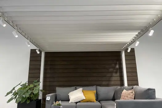 In showroom, sofa sits under Proteus White Aluminium pergola with open roof louvres, composite plank cladding on wall behind.