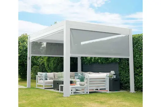 Proteus white aluminium pergola with 2 sides partially lowered stands over furniture on paved area in lawn next to hedge.