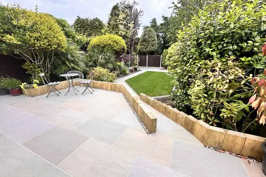 Raj Green Porcelain Paving forming a spacious patio area overlooking a well-kept garden with timber edging, mature shrubs, and a small bistro seating set.