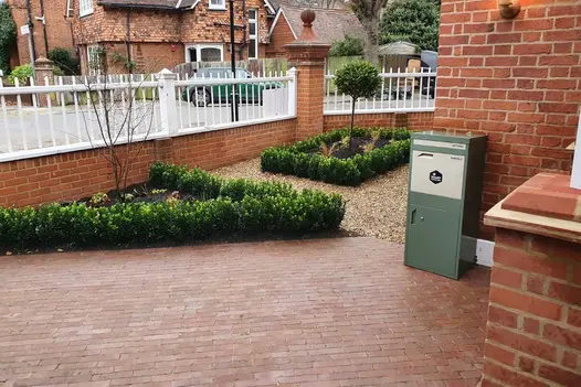 Roma Clay Brick Pavers in a warm, soft red tone laid in a neat stretcher bond pattern on a front driveway, bordered by low box hedging, gravel beds, and red-brick walls.