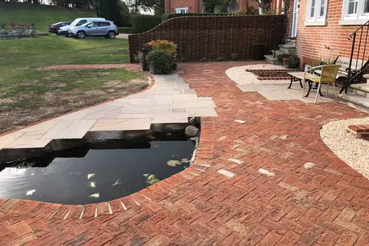 Romsey Antique Clay Brick Pavers in a warm, weathered red laid in a herringbone pattern around a garden pond, blending with cream stone paving and gravel features beside a traditional brick home.