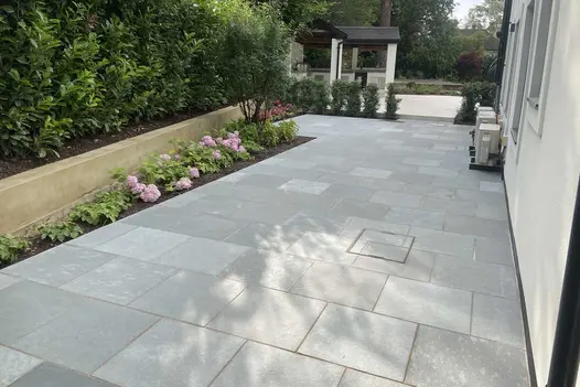 Kota Blue Limestone Paving laid in a contemporary patio beside raised planting beds and hydrangeas, leading toward a landscaped garden with mature trees.