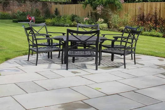 Tumbled Grey Sandstone Paving forming a curved patio with black metal dining furniture, set within a large, landscaped garden with lawn and planting borders.
