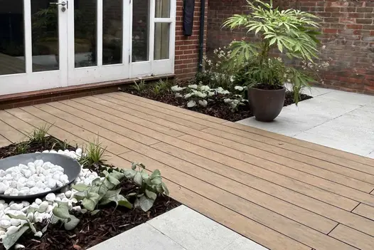Warm Teak Brushed Composite Decking forming a garden pathway surrounded by planting beds, white paving slabs, and a brick exterior wall.