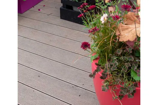 Warm Teak Grooved Composite Decking shown alongside a bright red planter filled with colourful flowers and foliage.