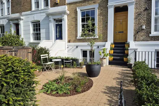 Westminster Clay Brick Pavers in a warm buff tone forming a front garden courtyard with planters, seating, and period terraced houses in the background.